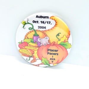 May include: A round, white button with a colorful illustration of a cornucopia filled with fruits and vegetables. The button has text that reads "Auburn Oct. 16/17, 2004" and "Placer Pacers I AVA V".