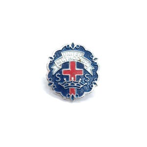 May include: A circular metal pin featuring a blue enamel background with a silver scalloped border. In the center, a red cross sits above a silver crown, flanked by the letter S on each side. A white banner across the top reads UNITED METHODIST.