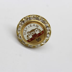 May include: Gold-coloured pin with a white enamel design featuring a lizard and the text "H&F.I. & A.W."  The pin is surrounded by clear rhinestones and has the number "50" in the centre.