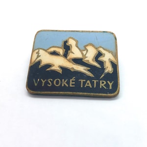 May include: A rectangular metal pin featuring a stylised mountain range graphic in cream against a dark blue base, set against a light blue sky. The text VYSOKÉ TATRY is printed in gold lettering along the bottom edge of the pin's face.