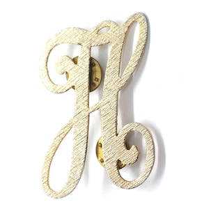 May include: Gold-coloured metal letter "H" pin with a textured surface. The pin features a script-style design with two pin backs. The letter is a warm gold colour and is set against a white background.