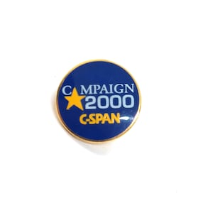 May include: A round pin with a blue background and gold trim. The pin features the words "CAMPAIGN 2000" in white and the word "C-SPAN" in yellow. A gold star is also present.