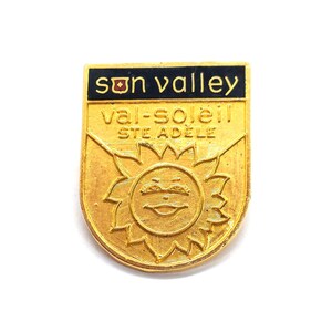 May include: A gold-colored metal pin with the words "Sun Valley" in black lettering at the top. Below, it reads "val-soleil STE ADELE" in gold. The pin features a smiling sun design.