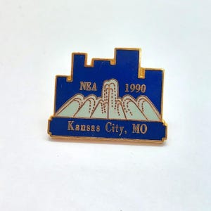 May include: A vintage pin with a blue skyline design, gold border, and the text "NEA 1990" and "Kansas City, MO". The pin also features a light blue mountain range design.