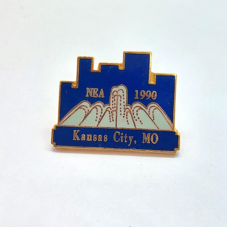 May include: A vintage pin with a blue skyline design, gold border, and the text "NEA 1990" and "Kansas City, MO". The pin also features a light blue mountain range design.
