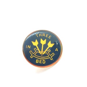 May include: A circular navy blue badge with gold lettering and imagery. The text reads "THREE IN A BED" with three gold arrows. The badge has a gold edge.