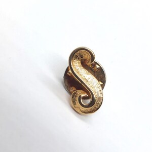 May include: A gold-toned metal lapel pin shaped like the letter S. The surface of the letter features a textured, etched finish with a polished edge. The pin includes a circular backing mechanism visible behind the letter, designed for attachment to fabric.