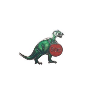 May include: A green dinosaur pin with a red circle that has the number 500 on it. The pin is made of metal and has a glossy finish. The dinosaur is standing on its hind legs.