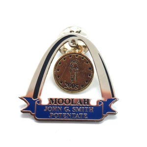 May include: A silver-toned pin with a blue banner that reads "MOOLAH JOHN G. SMITH POTENTATE." A bronze-colored medallion with a figure and the year 2005 hangs from the pin. The pin has a silver arch design.
