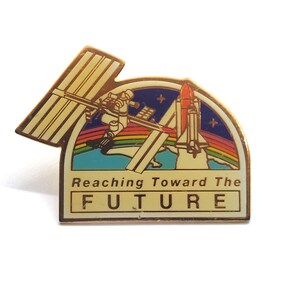 May include: A vintage pin featuring a space shuttle, astronaut, and the International Space Station. The pin has a gold border and the text "Reaching Toward The FUTURE" on a rectangular base. The background includes a rainbow and stars.