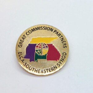 May include: A gold-coloured pin with the text "ELCA-SOUTHEASTERN SYNOD GREAT COMMISSION PARTNERS" around the edge. The centre of the pin features a stylised outline of the state of Tennessee with a globe in the centre. The globe is coloured in red, yellow, green, and purple.