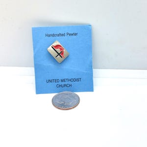 May include: A handcrafted pewter pin with a red and black cross design on a silver background. The pin is on a blue card with the text "Handcrafted Pewter" and "UNITED METHODIST CHURCH". A coin is visible below the card.