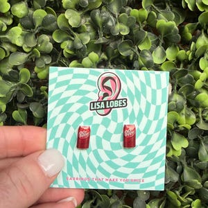 May include: A pair of red Dr. Pepper can earrings on a teal and white checkered card. The card has the text "LISA LOBES" and "EARRINGS THAT MAKE YOU SMILE". The earrings are displayed against a backdrop of green foliage.