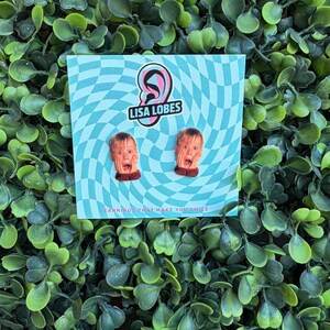 Pu&ograve; includere: Un paio di orecchini originali con un design a volto sorpreso. Gli orecchini sono esposti su una carta a scacchi blu e bianca con il testo "LISA LOBES" e "EARRINGS THAT MAKE YOU SMILE". Lo sfondo &egrave; una fitta disposizione di foglie verdi.