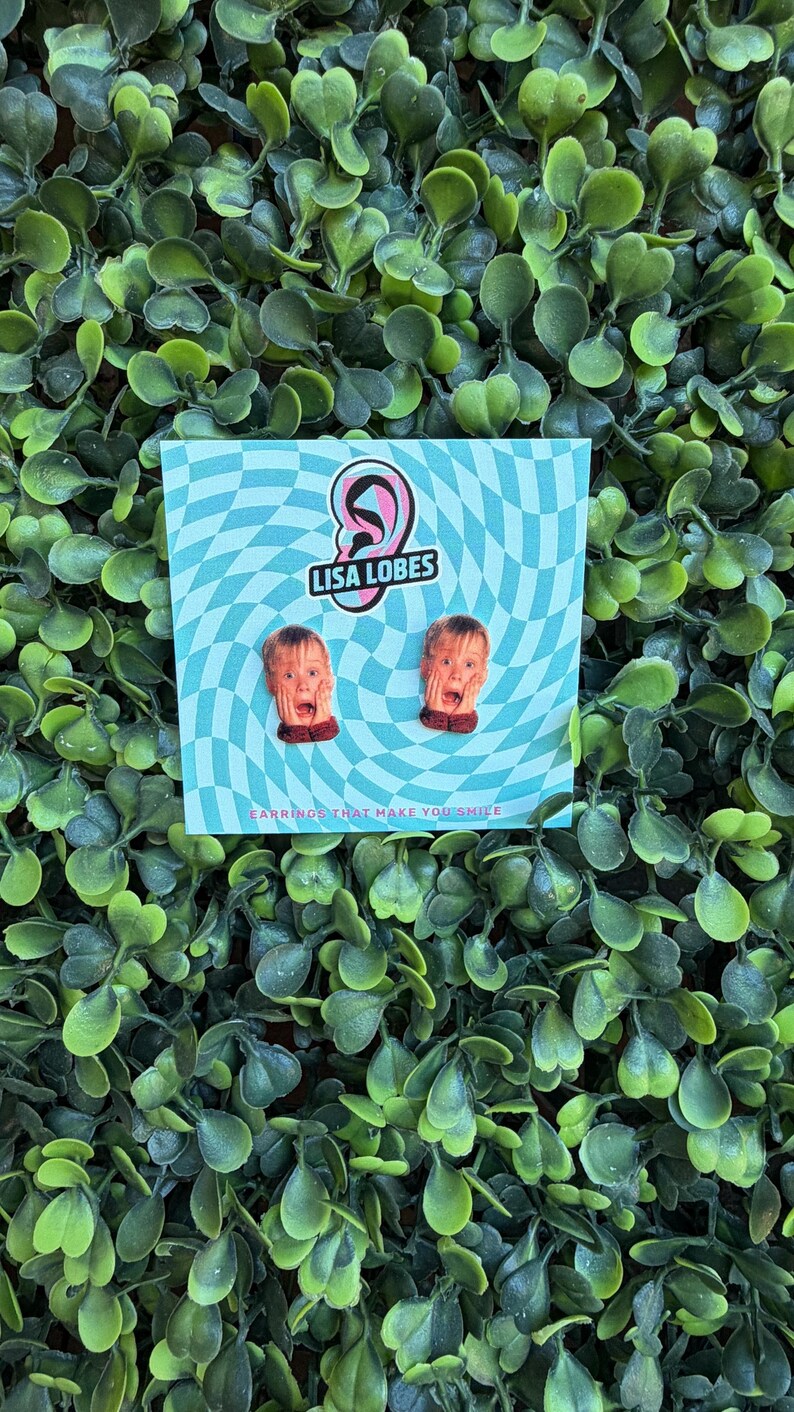 Pu&ograve; includere: Un paio di orecchini originali con un design a volto sorpreso. Gli orecchini sono esposti su una carta a scacchi blu e bianca con il testo "LISA LOBES" e "EARRINGS THAT MAKE YOU SMILE". Lo sfondo &egrave; una fitta disposizione di foglie verdi.