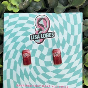 May include: A pair of Dr. Pepper can earrings on a teal and white checkered background. The earrings are red with white text. The card has the text "Lisa Lobes" and "Earrings that make you smile".