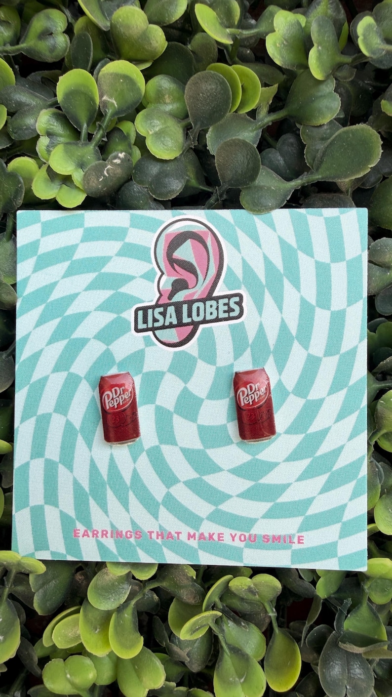 May include: A pair of Dr. Pepper can earrings on a teal and white checkered background. The earrings are red with white text. The card has the text "Lisa Lobes" and "Earrings that make you smile".