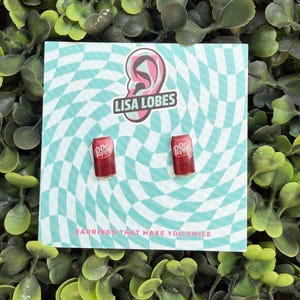 May include: A pair of red Dr. Pepper can earrings on a teal and white checkered background. The earrings are on a card with the text "LISA LOBES" and "EARRINGS THAT MAKE YOU SMILE". The background is surrounded by green foliage.