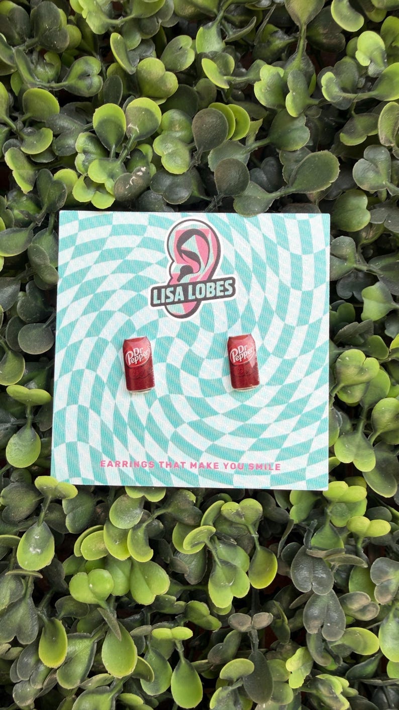 May include: A pair of red Dr. Pepper can earrings on a teal and white checkered background. The earrings are on a card with the text "LISA LOBES" and "EARRINGS THAT MAKE YOU SMILE". The background is surrounded by green foliage.