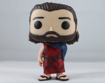 May include: A vinyl collectible figure featuring a character with long brown hair and a full beard. The figure wears a red kilt with a black crisscross pattern, a dark blue shoulder wrap, a black belt, and simple black sandals on its feet.