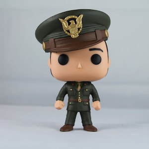 May include: A vinyl figure of a man in a military uniform. The figure wears a dark green uniform with a brown belt and shoes. A dark green hat with a gold emblem sits atop the figure's head. The figure has black eyes and a neutral expression.