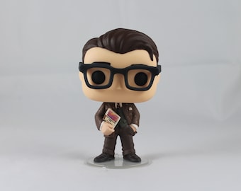 May include: A brown and black pop vinyl figure of a man wearing a brown suit, a white shirt, and a black tie. He is wearing black framed glasses and holding a small book in his right hand. The figure is standing on a clear plastic base.