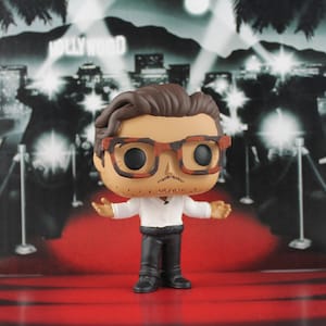 Peut inclure: Une figurine Funko Pop en vinyle d'un homme portant des lunettes, une chemise blanche, un pantalon noir et des chaussures noires. La figurine est sur un tapis rouge avec une toile de fond Hollywood.