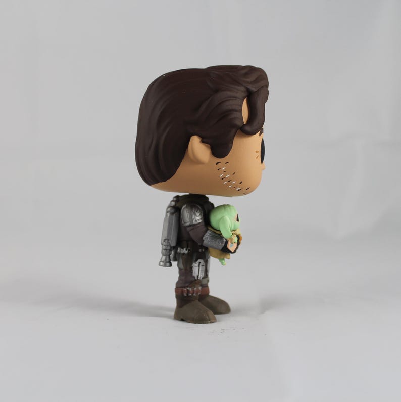 May include: A Funko Pop vinyl figure of The Mandalorian holding Grogu, also known as Baby Yoda. The figure features a brown-haired character in silver and brown armor, with Grogu in a green robe. The figure is standing on a white surface.