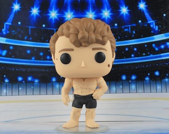May include: A vinyl collectible figure featuring a person with curly brown hair, a large head, and black circular eyes. The figure has a light skin tone, a bare chest with small brown spots, and is wearing black athletic shorts.