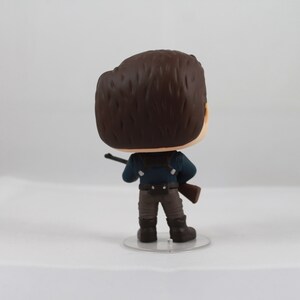 May include: A vinyl figure of a character from a movie or TV show, viewed from the back. The figure has brown hair, a blue shirt, brown pants, and black boots. It is holding a rifle and standing on a clear plastic base.