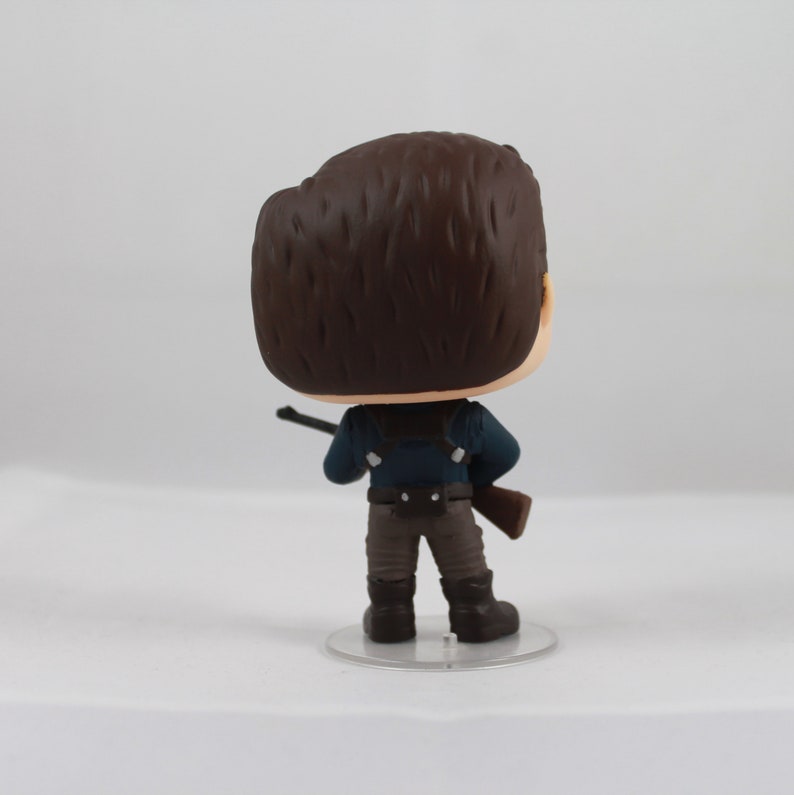 May include: A vinyl figure of a character from a movie or TV show, viewed from the back. The figure has brown hair, a blue shirt, brown pants, and black boots. It is holding a rifle and standing on a clear plastic base.