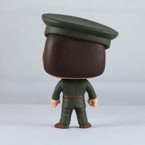 Peut inclure: Figurine en vinyle d'une personne en uniforme militaire, vue de dos. La figurine porte un uniforme vert fonc&eacute;, une casquette assortie et des chaussures marron. La figurine a les cheveux bruns et une ceinture marron.