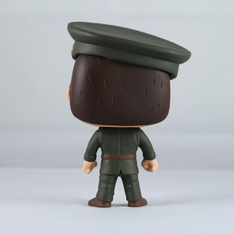 Peut inclure: Figurine en vinyle d'une personne en uniforme militaire, vue de dos. La figurine porte un uniforme vert fonc&eacute;, une casquette assortie et des chaussures marron. La figurine a les cheveux bruns et une ceinture marron.