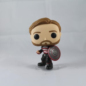 May include: A Funko Pop vinyl figure of a character with brown hair, a beard, and a red, white, and black costume. The figure is holding a shield with red and silver details. The figure is standing on a clear plastic base.