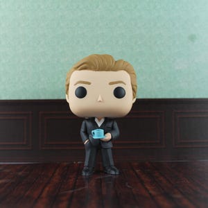 May include: A vinyl collectible figure featuring a character with light brown hair, wearing a dark grey suit with a white shirt underneath. The figure has large black eyes and is holding a small, light blue teacup and saucer in its hands.