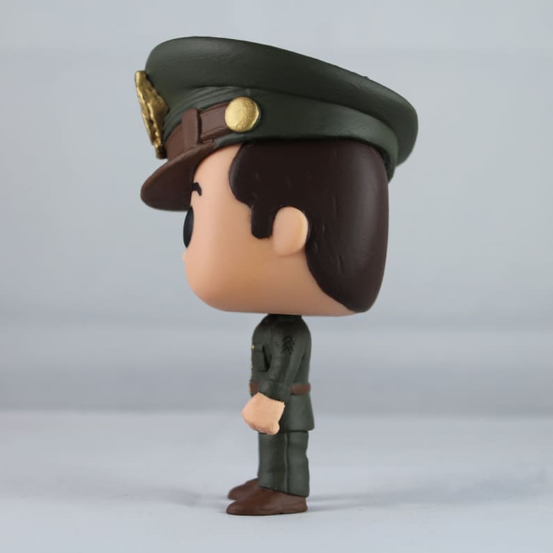 Peut inclure: Vue lat&eacute;rale d'une figurine de collection repr&eacute;sentant un homme en uniforme militaire. La figurine a un chapeau vert fonc&eacute; avec un embl&egrave;me dor&eacute; et un bord marron. L'uniforme est vert fonc&eacute; avec une ceinture marron et des chaussures marron.