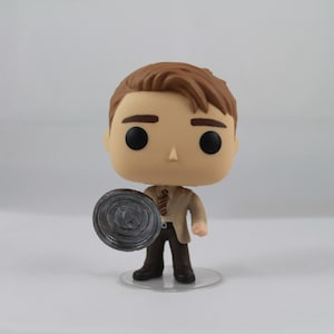 May include: A Funko Pop vinyl figure of a character with brown hair, wearing a tan suit, brown trousers, and a striped tie. The figure is holding a grey, circular object. The figure is standing on a clear plastic base.