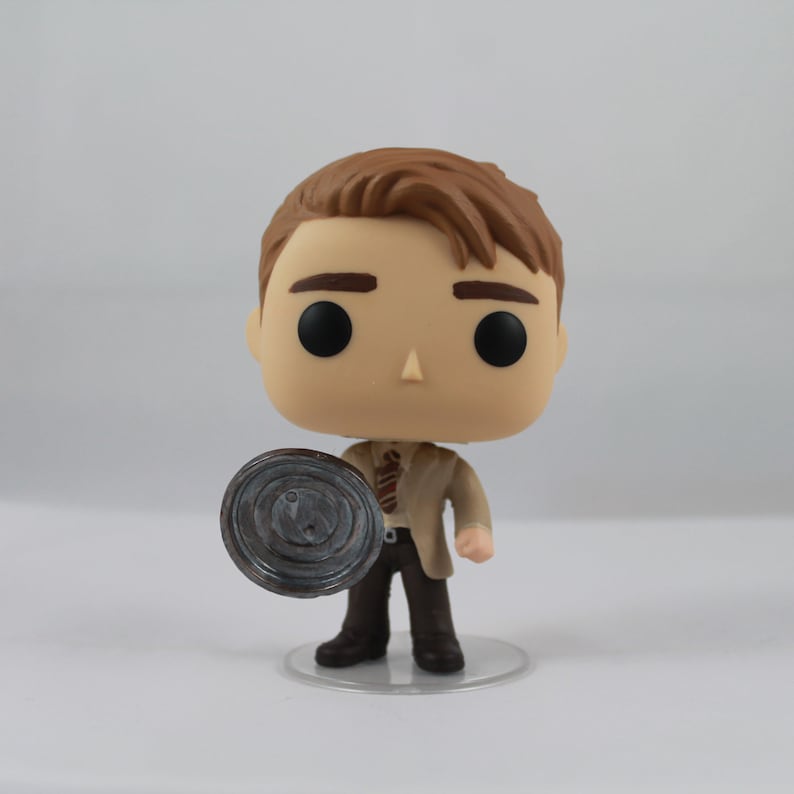 May include: A Funko Pop vinyl figure of a character with brown hair, wearing a tan suit, brown trousers, and a striped tie. The figure is holding a grey, circular object. The figure is standing on a clear plastic base.