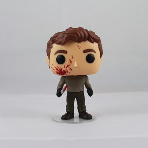 May include: A vinyl figure of a character with brown hair and a bloodied face. The figure wears a brown shirt, dark pants, and black gloves, holding a small knife. The figure stands on a clear plastic base.