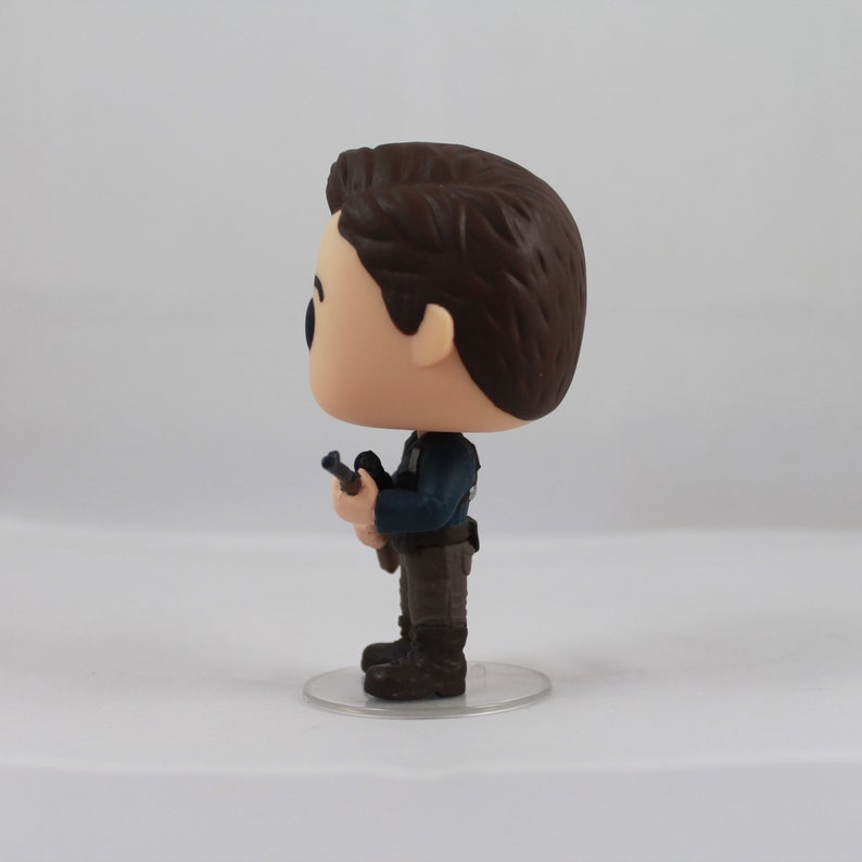 May include: A vinyl figure of a man in a dark blue jacket, brown pants, and black boots, holding a gun. The figure has brown hair and is standing on a clear, circular base. The figure is a collectible item.