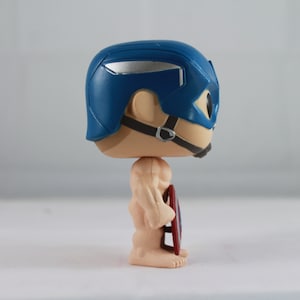 May include: A vinyl figurine of a character wearing a blue helmet with silver accents and a black chin strap. The figure has a light skin tone, a muscular build, and is holding a circular red, white, and blue shield at its side in a profile view.