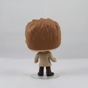 May include: A vinyl figure of a person with brown hair, wearing a beige coat and dark brown boots. The figure is standing on a clear, circular base. The back of the figure is facing the camera.