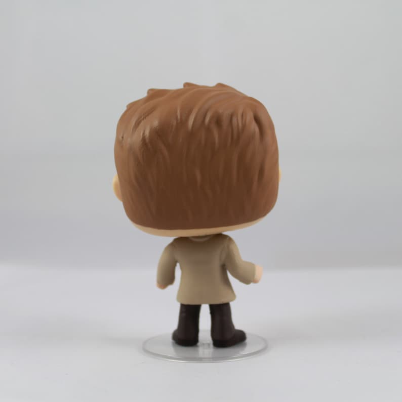 May include: A vinyl figure of a person with brown hair, wearing a beige coat and dark brown boots. The figure is standing on a clear, circular base. The back of the figure is facing the camera.