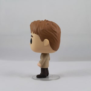 May include: A Funko Pop vinyl figure of a character with light skin, brown hair, and a tan coat. The figure stands on a clear plastic base. The figure's eyes are black, and it has a small nose and mouth.