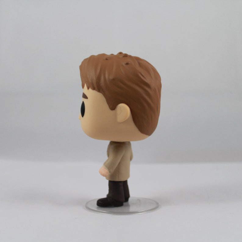 May include: A Funko Pop vinyl figure of a character with light skin, brown hair, and a tan coat. The figure stands on a clear plastic base. The figure's eyes are black, and it has a small nose and mouth.