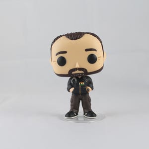 May include: A Funko Pop vinyl figure of a man with a beard, wearing a black jacket and brown trousers. The figure has a small Indian flag on the jacket and is standing on a clear plastic base. The figure has a large head and black eyes.