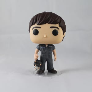 May include: A Funko Pop vinyl figure of a character in a gray jumpsuit, holding a small, black and white object. The figure has brown hair, large black eyes, and is standing on a clear plastic base. The jumpsuit has a small patch.