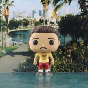 May include: A vinyl collectible figure featuring a character with brown hair, a mustache, and light stubble. The figure wears a yellow t-shirt with a graphic print, red shorts, white socks, and grey sneakers, standing on a flat surface.