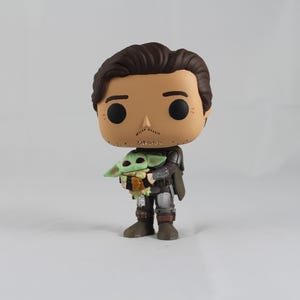 May include: A vinyl figure of a character from a popular science fiction series. The figure depicts a man in a dark outfit holding a small green creature. The figure has a large head and black eyes, with brown hair and a mustache.