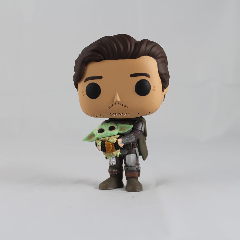 May include: A vinyl figure of a character from a popular science fiction series. The figure depicts a man in a dark outfit holding a small green creature. The figure has a large head and black eyes, with brown hair and a mustache.